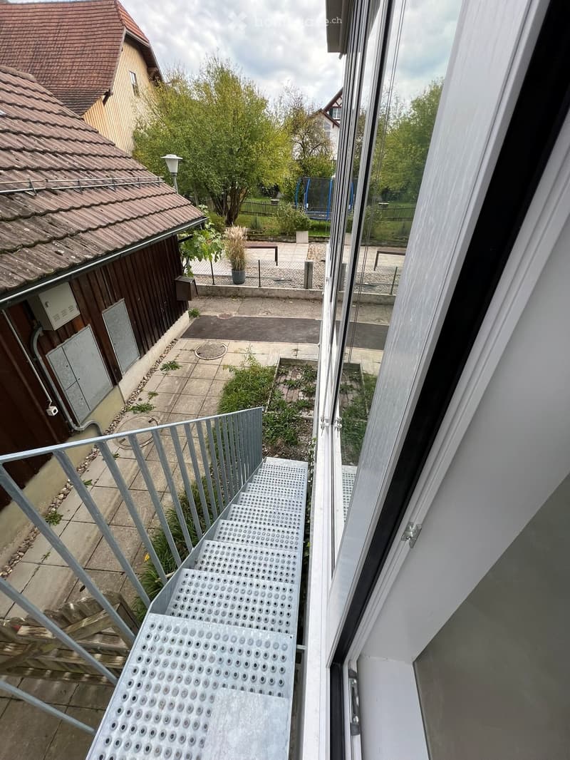 Balcony with metal stairs leading down to a small outdoor area with concrete flooring, greenery, and a wooden shed.