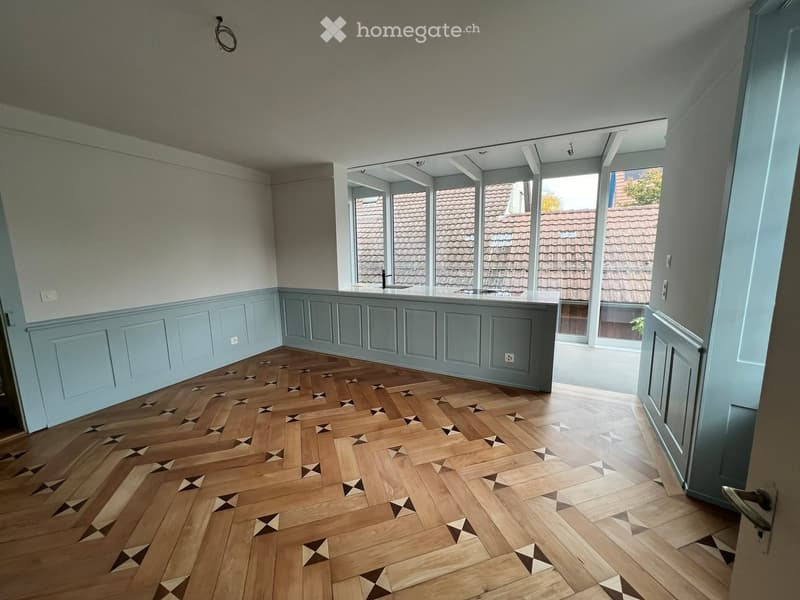 Empty room with light blue walls, wooden herringbone floor, and large windows overlooking a tiled roof.