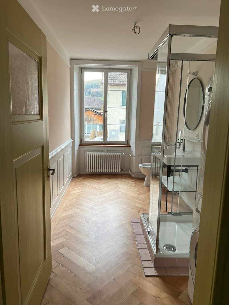 Bathroom with herringbone wooden flooring, glass-enclosed shower, toilet, sink, and mirror. Features a large window with a view of the outside.