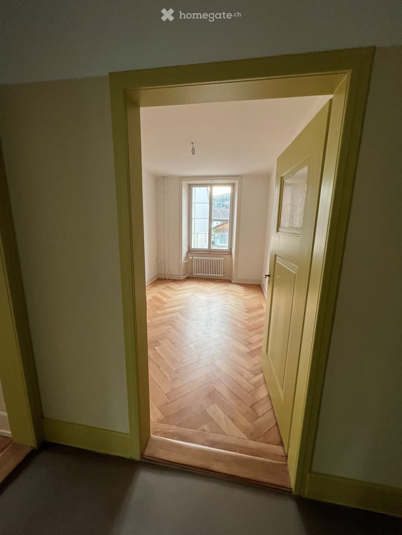 Empty room with wooden herringbone flooring, white walls, a window with a radiator below, and a partially open door leading to another room.