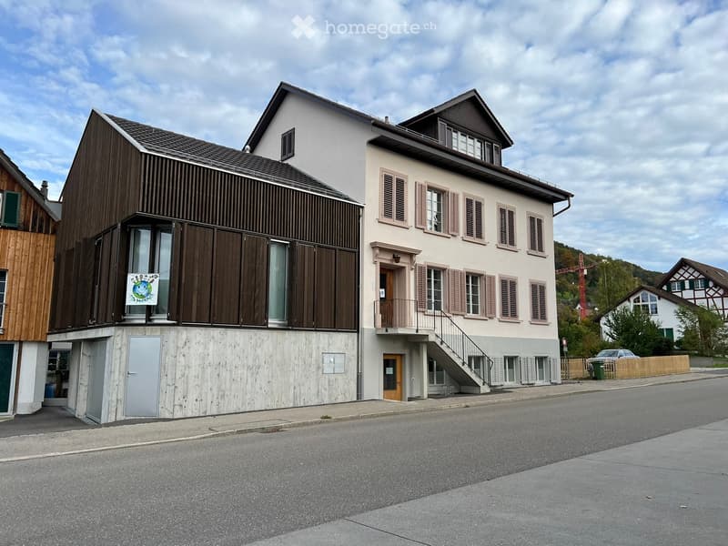 Two-story residential building with a modern and traditional design, featuring a staircase leading to a balcony, multiple windows with shutters, and a mix of wood and plaster exterior finishes.