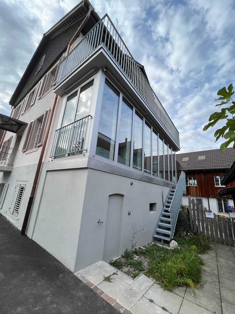 Modern two-story house with white exterior, large windows, and a balcony on the upper floor. Features a staircase leading to the entrance and a small garden area.