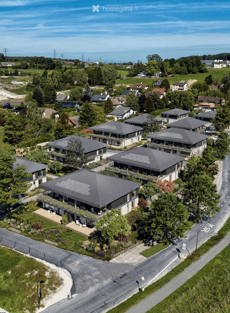 Aerial view of a modern residential complex with multiple houses featuring grey roofs and solar panels, surrounded by greenery and trees.