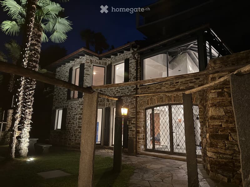 Two-story stone house with large windows and a wooden pergola, surrounded by palm trees and a stone-paved patio.