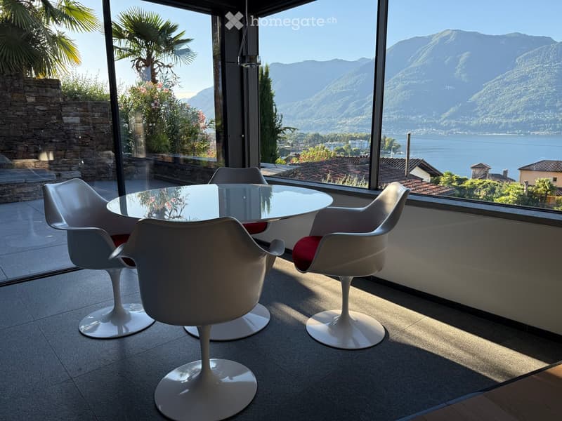 Modern dining area with a round glass table and four white chairs, overlooking a scenic lake and mountain view.