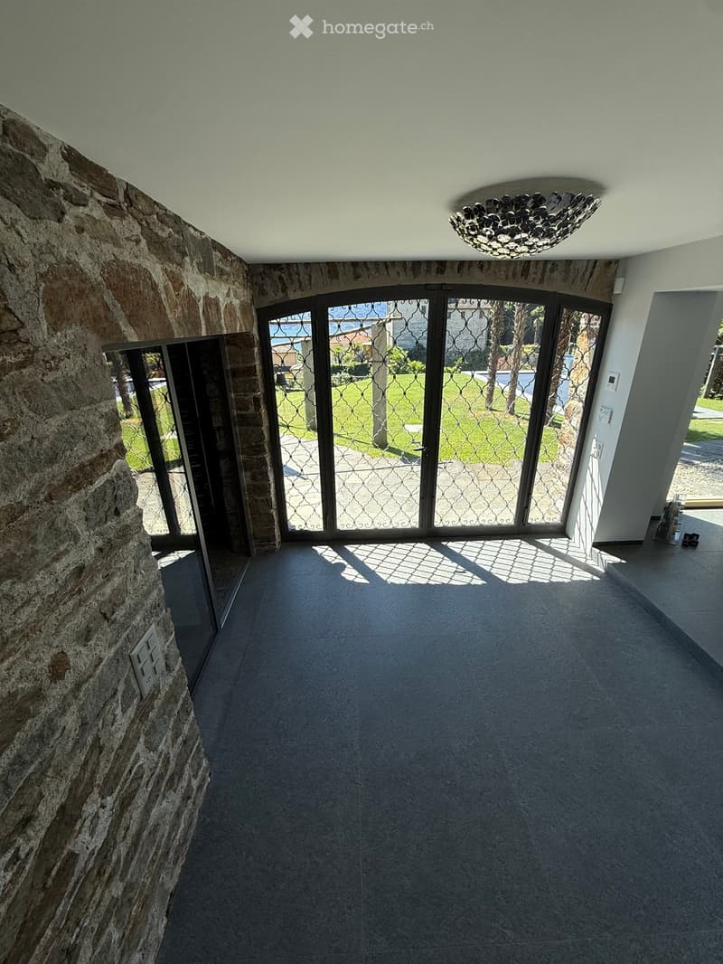 Empty hallway with stone walls, tiled floor, large glass doors leading to a garden, and a decorative ceiling light fixture.