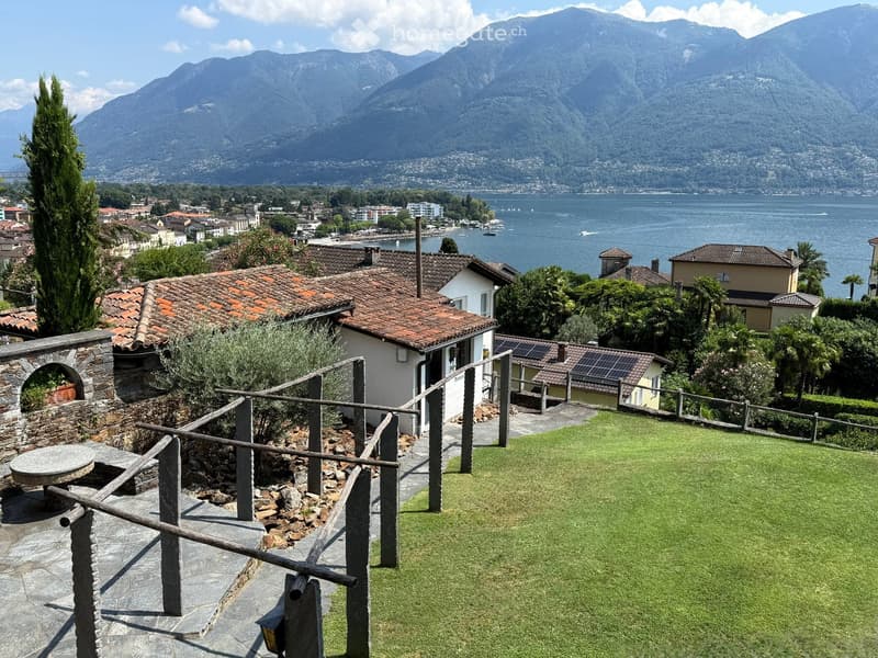 A scenic garden area with a stone wall, wooden railing, and a round stone table, overlooking a lake and mountains.