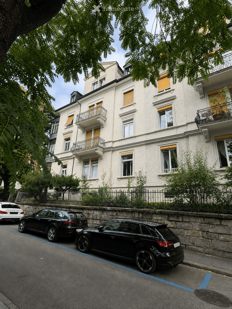 A multi-story residential building with white facade, multiple windows, and balconies. The building is surrounded by greenery and has a stone fence along the sidewalk. Several cars are parked on the street in front of the building.