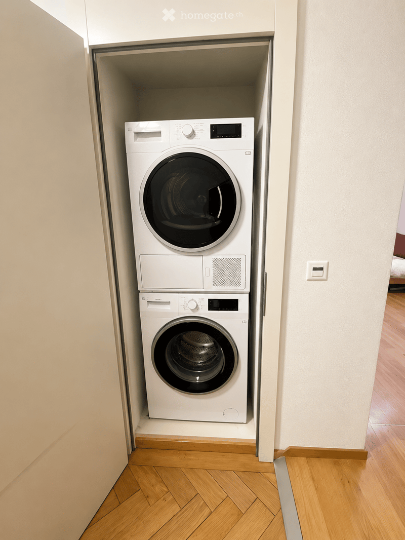 A compact bathroom with a stacked washing machine and dryer unit, white walls, and wooden flooring.