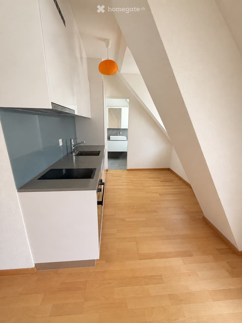 Empty attic space with wooden flooring, white walls, and a small kitchen area featuring a sink, faucet, and cabinets. A hallway leads to a small bathroom with a sink and mirror.