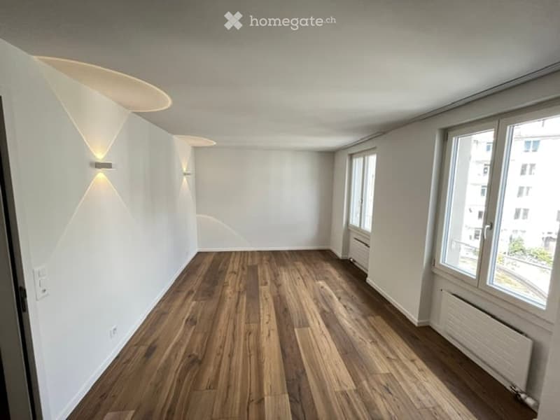 Empty room with wooden flooring, white walls, large windows, and modern wall-mounted lighting fixtures.