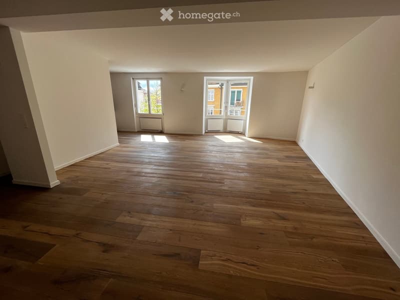 Empty room with wooden flooring, white walls, and large windows allowing natural light. Radiators are visible under the windows.