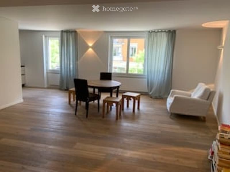 Bright living room with wooden flooring, large window with grey curtains, white sofa, dining table with chairs, and two small stools.