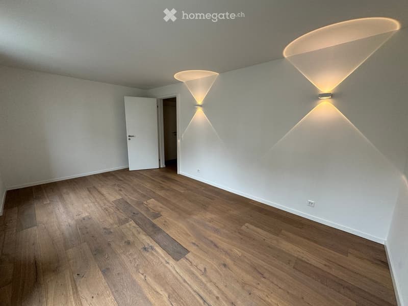 Empty room with wooden flooring, white walls, and modern wall-mounted lighting fixtures. A door is visible on the left side.