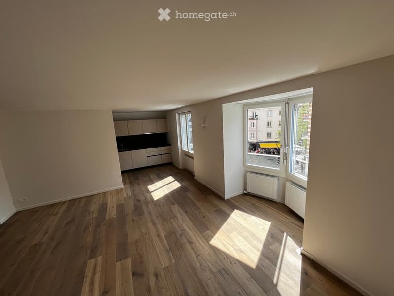 Empty room with wooden flooring, white walls, and large windows allowing natural light to enter. The room features a small kitchen area with cabinets and a countertop visible in the background.