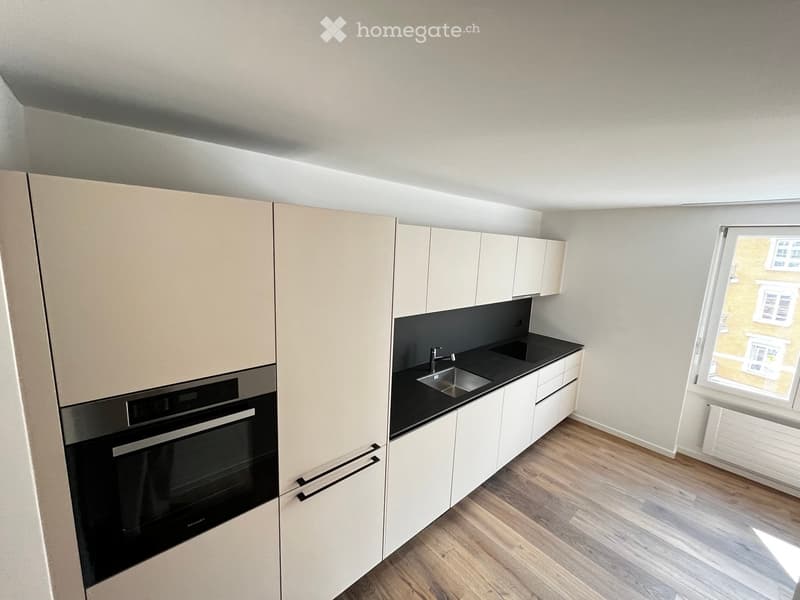 Modern kitchen with white cabinets, black countertop, stainless steel sink, and built-in oven. Light wooden flooring and large window.