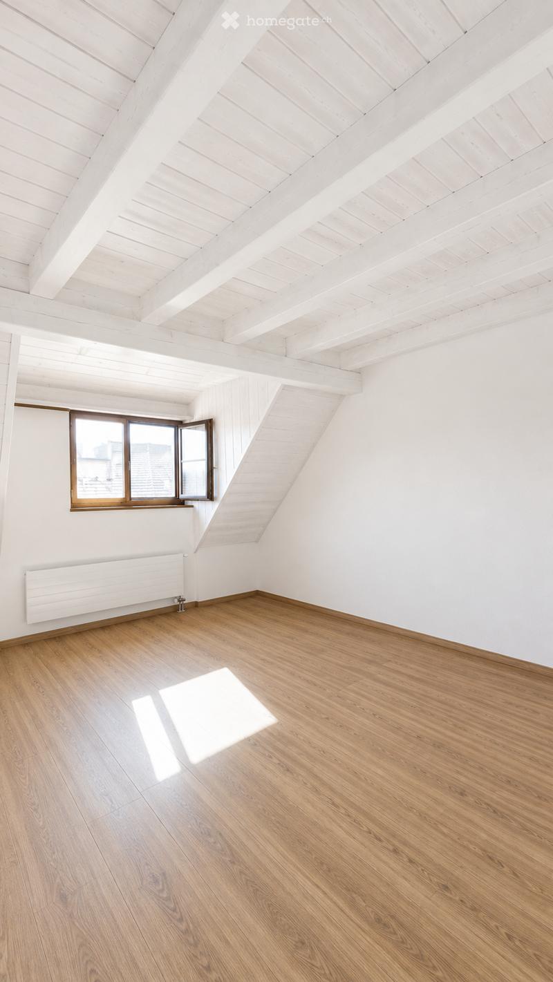 Empty attic room with wooden floors, white walls, exposed wooden beams, and a single window with wooden frame.