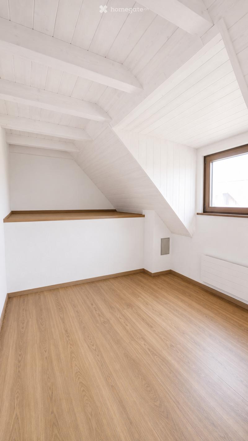 Empty attic room with wooden flooring, white walls, and exposed wooden beams on the ceiling. Features a single window with wooden frame and a built-in wooden shelf.