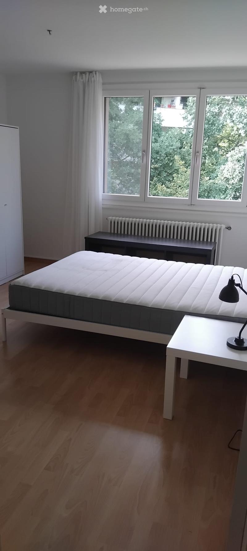 Minimalist bedroom with a white bed, small white table with a black lamp, bench, radiator, and large windows with white curtains.