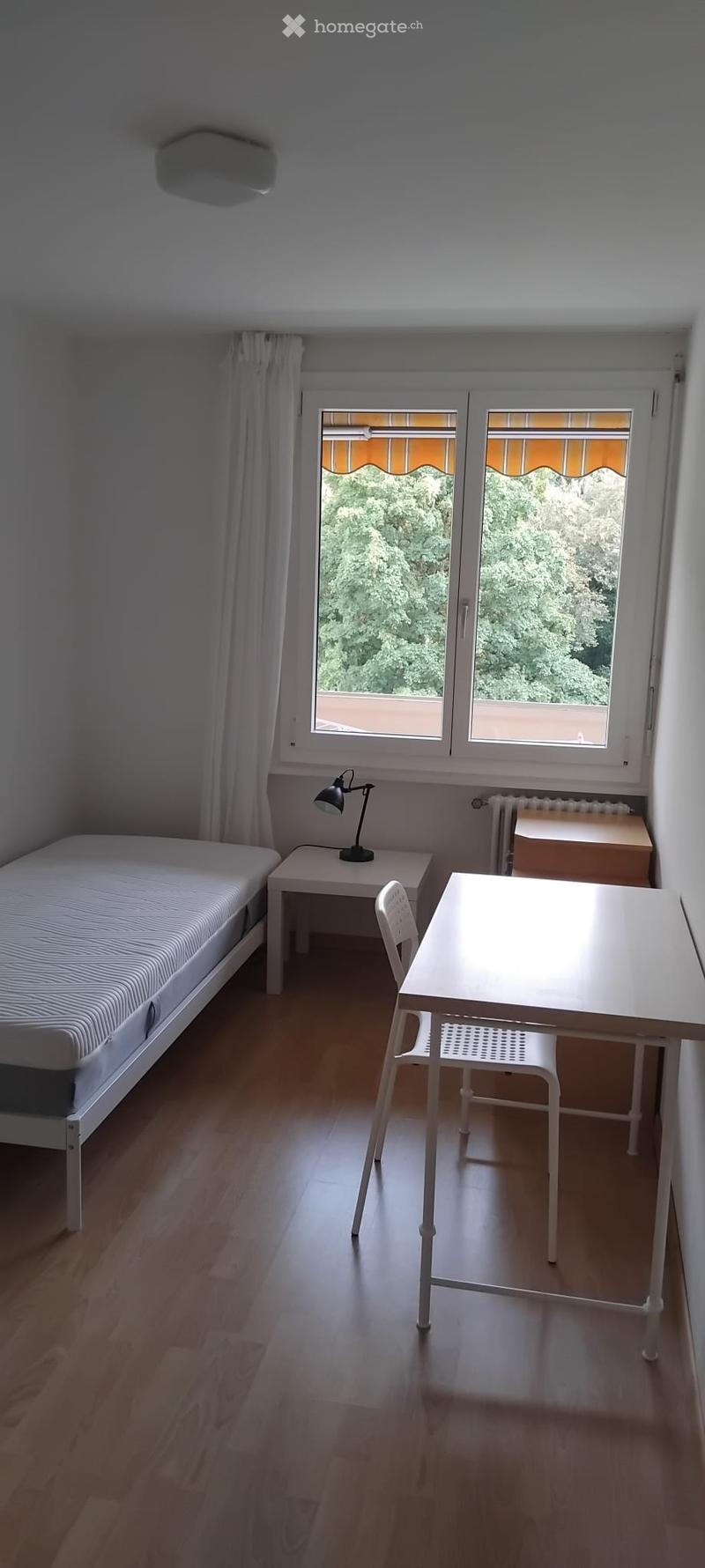 A small, well-lit bedroom with a single bed, a desk, a chair, and a lamp. The room has wooden flooring and large windows with white curtains, offering a view of greenery outside.