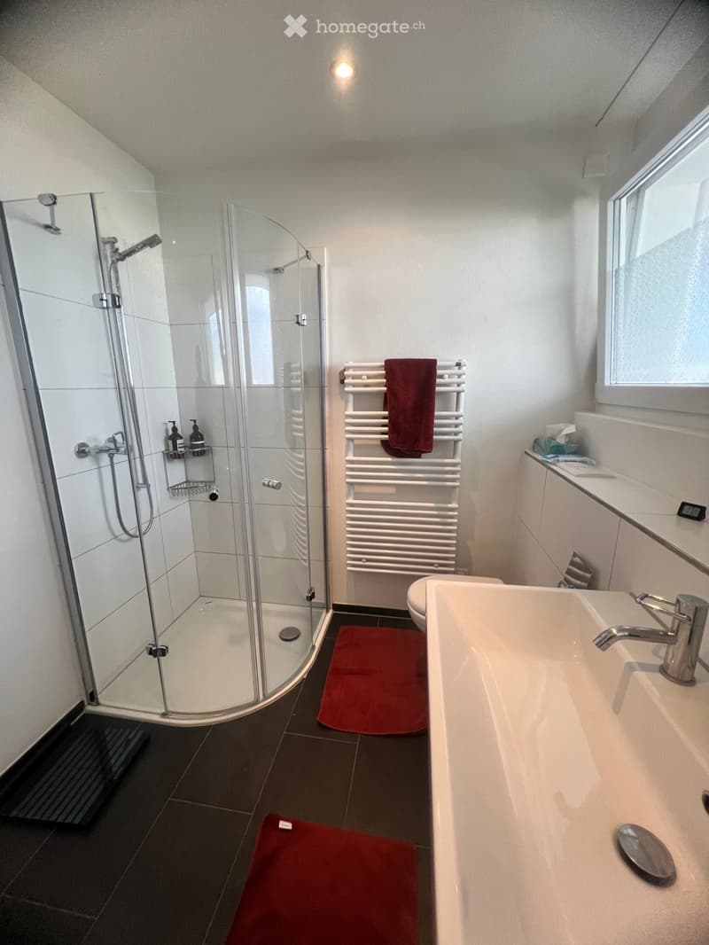 A shower cubicle, glass walk-in shower, shower head, shower hose, shower rack, toilet, sink, white tile flooring, window with blinds, towel rack with a red towel, and other bathroom amenities on the counter.