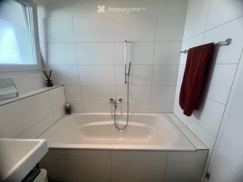 modern white tiled walls, large bathtub, separate sink, shower head, towel rack with red towel, mirror on the wall, green bottle on the counter, floor without visible flooring