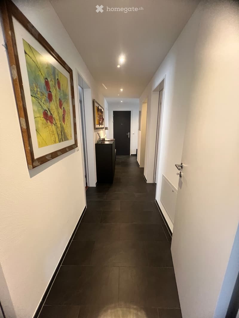 An apartment hallway with black tile flooring, white walls, and illuminated ceilings. The hallway is adorned with framed artwork on the wall and a black cabinet with a lamp in the distance. A few doors are situated on either side of the hallway.