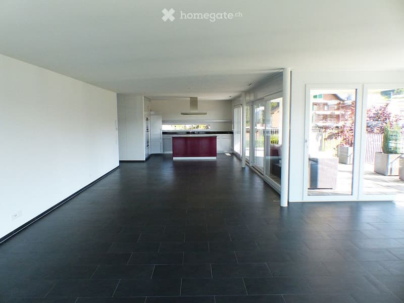 Empty room with white walls, black tile flooring, kitchen area with a red counter, a refrigerator and an extractor hood, sliding glass doors leading to a balcony with plants