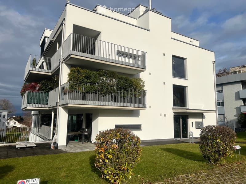 Modern white apartment building with multiple balconies, raised ground floor, and well-maintained green lawn in front.