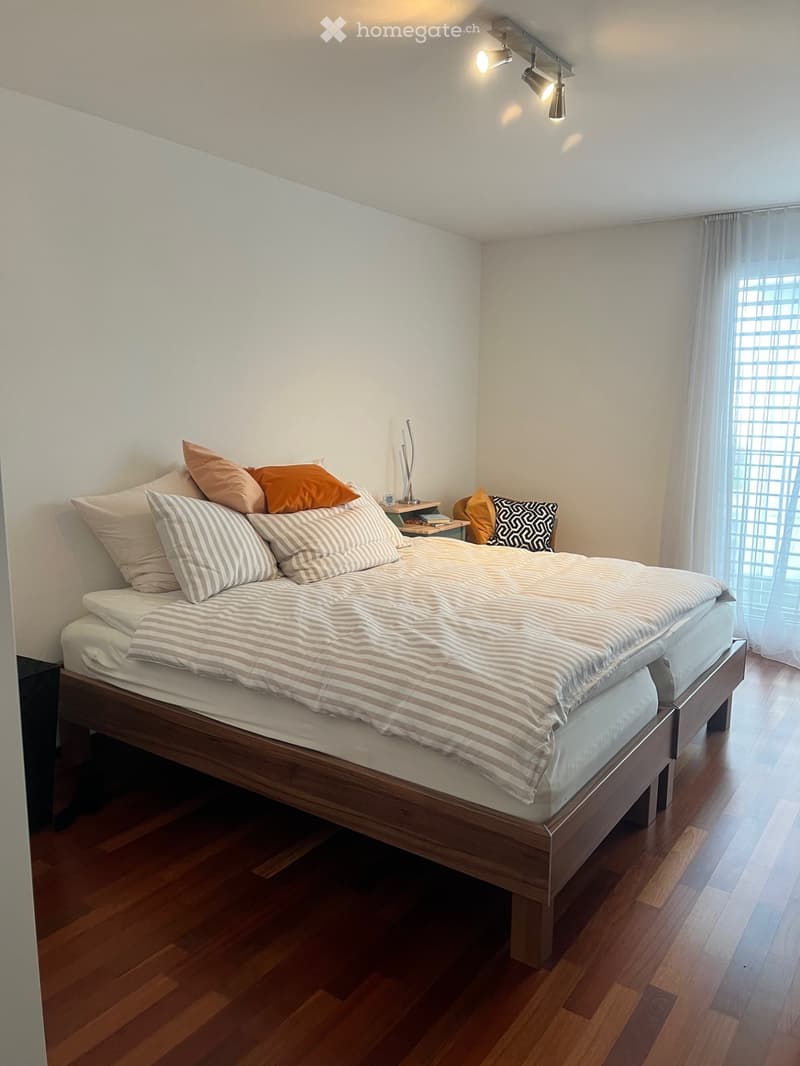A bedroom with a double bed, wooden headboard, white walls, hardwood floors, two nightstands, pillows, white bedding, window with white curtains, and a nightstand with a lamp.