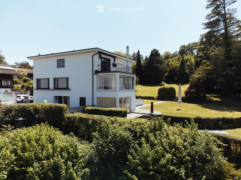 A two-story white house with large windows and a balcony, surrounded by a well-maintained garden and trees.