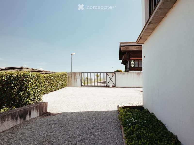 A gated driveway leads to a house with a balcony and a wooden structure, surrounded by neatly trimmed hedges and small plants.