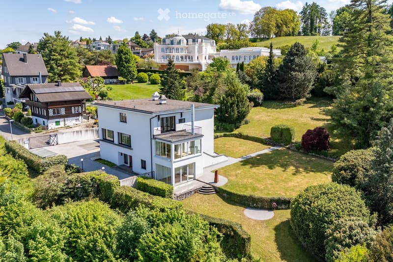 Aerial view of a modern white house with a balcony, surrounded by a well-maintained lawn and trees, with neighboring houses and a scenic hillside in the background.