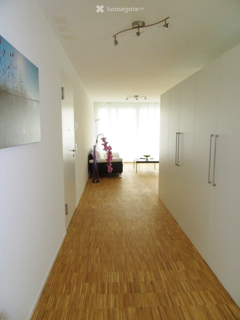 A modern hallway with wooden flooring, white walls, and recessed lighting. Features a large painting on the left wall and a small seating area visible at the end.