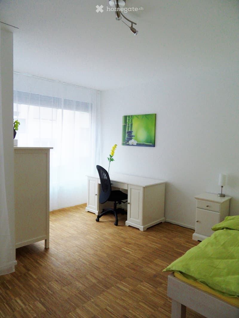 Bright bedroom with wooden flooring, a bed with green bedding, a desk with a chair, a nightstand with a lamp, and a painting on the wall.