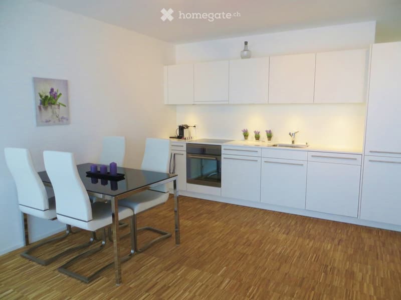 Modern kitchen and dining area with white cabinets, a black dining table with four white chairs, and a built-in oven. The floor is wooden, and there is a small sink with a faucet and a pendant light above the cabinets.
