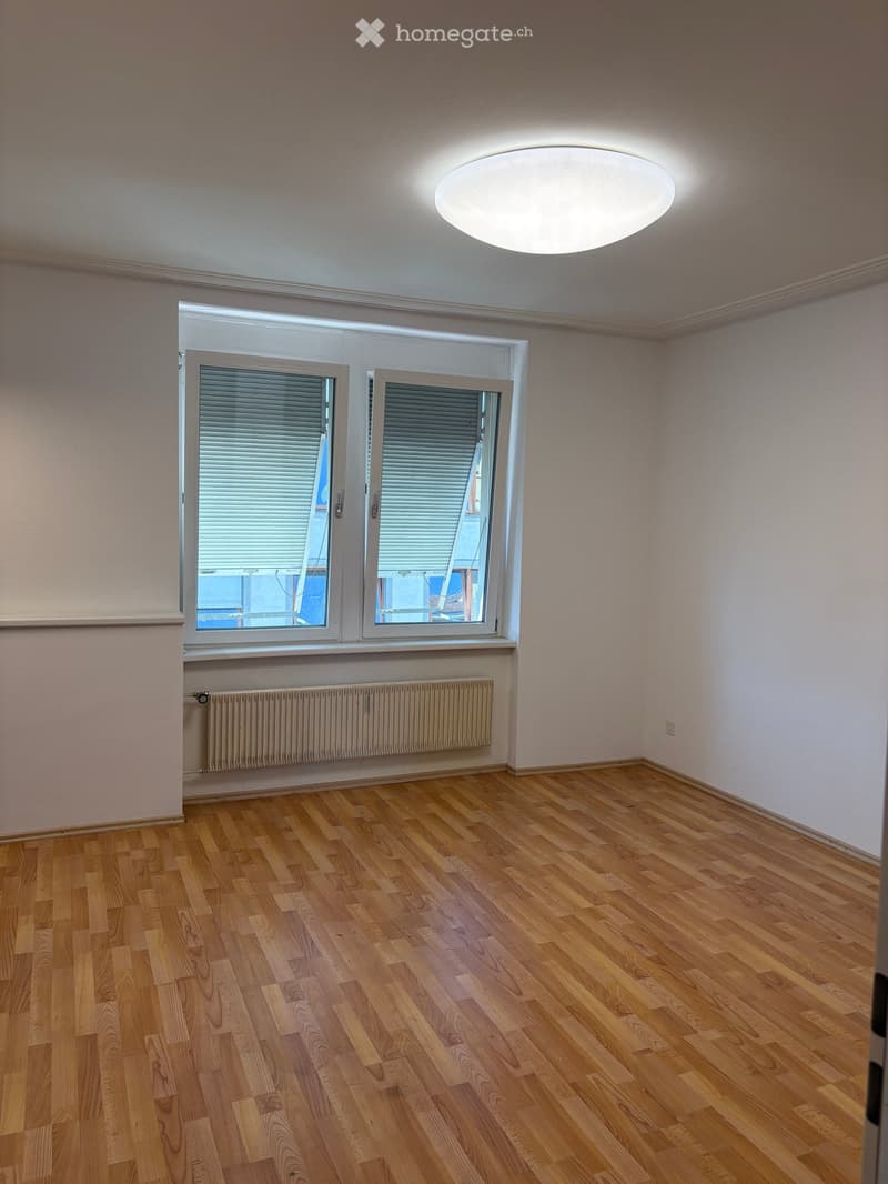 Empty room with wooden flooring, large windows with blinds, and a ceiling light fixture.
