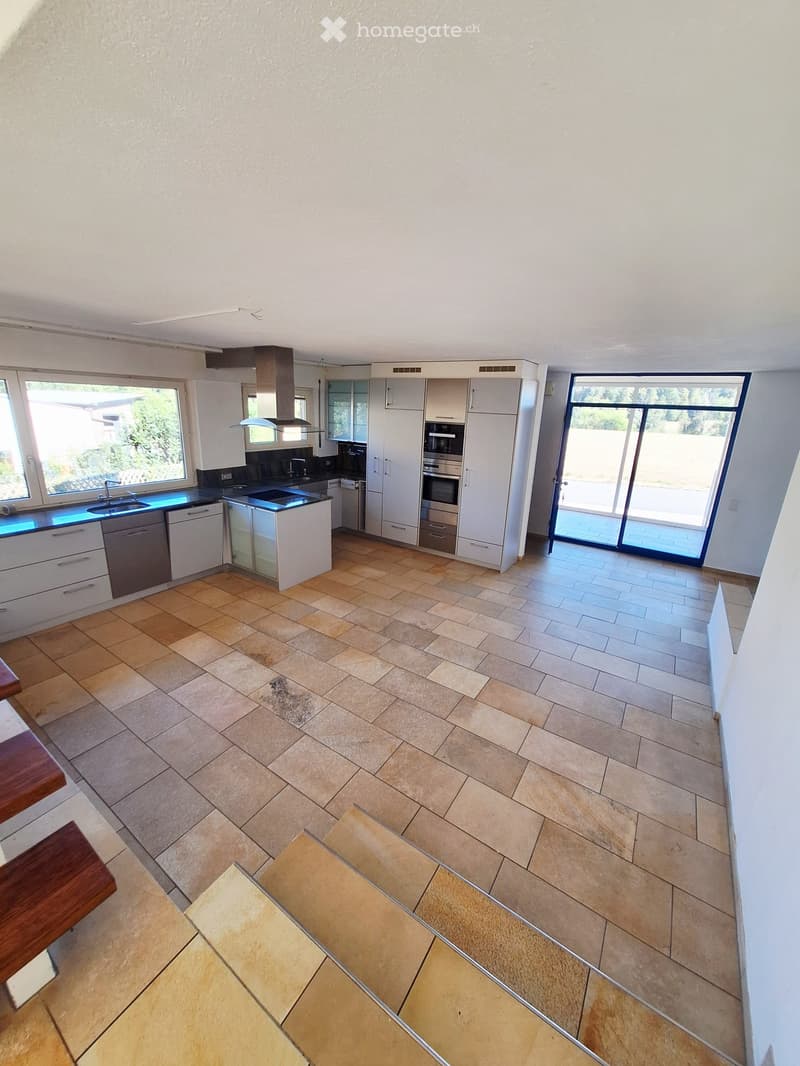 Modern kitchen with white cabinets, stainless steel appliances, and large windows. Features a central island with a sink and a sliding glass door leading to an outdoor area.