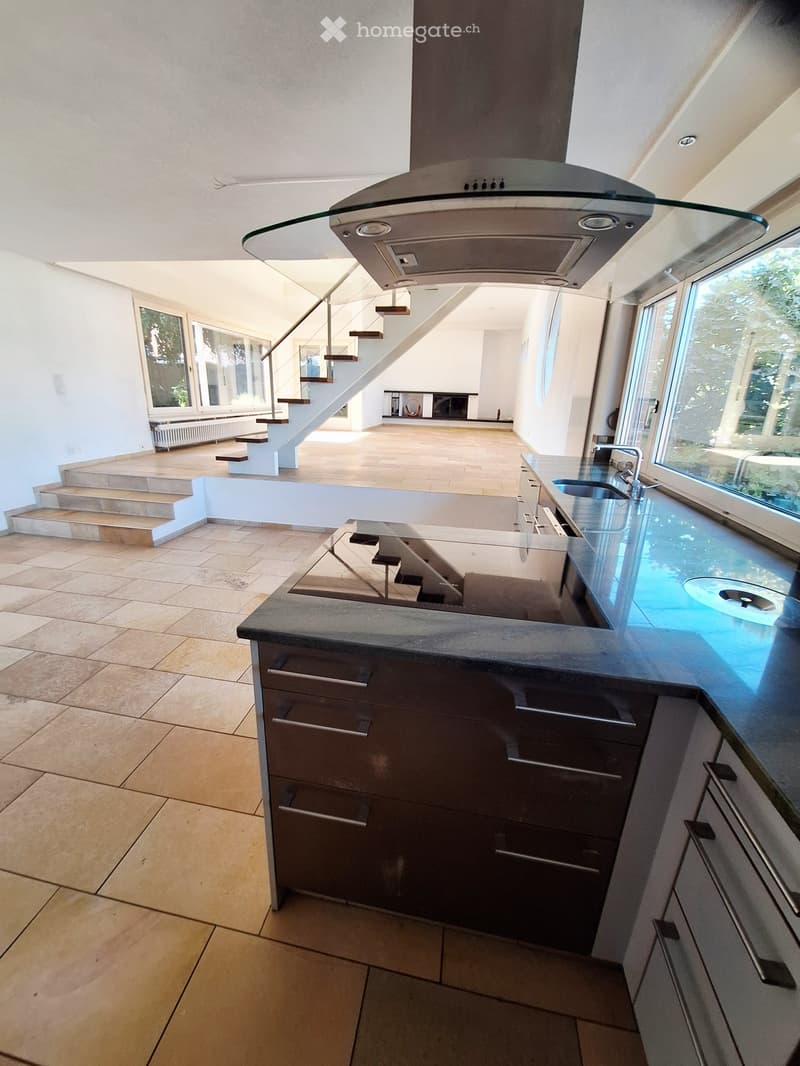Modern kitchen with dark cabinets, a central island, and a range hood. The space features large windows and a staircase leading to another level.