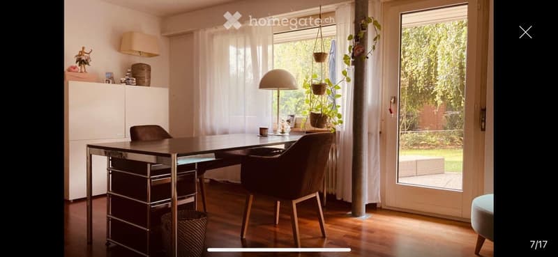 Modern dining area with a sleek black table, two dark chairs, a white cabinet, and a hanging lamp. Large windows with sheer curtains let in natural light, and a potted plant adds a touch of greenery.