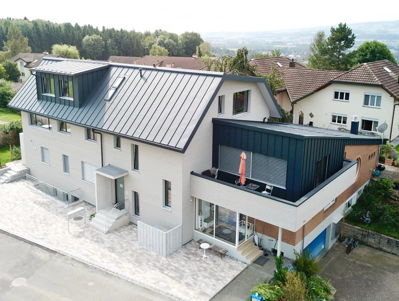 Modern two-story house with grey metal roof, white walls, and dark grey accents. Features include a raised ground floor, balcony, patio, and outdoor seating area.