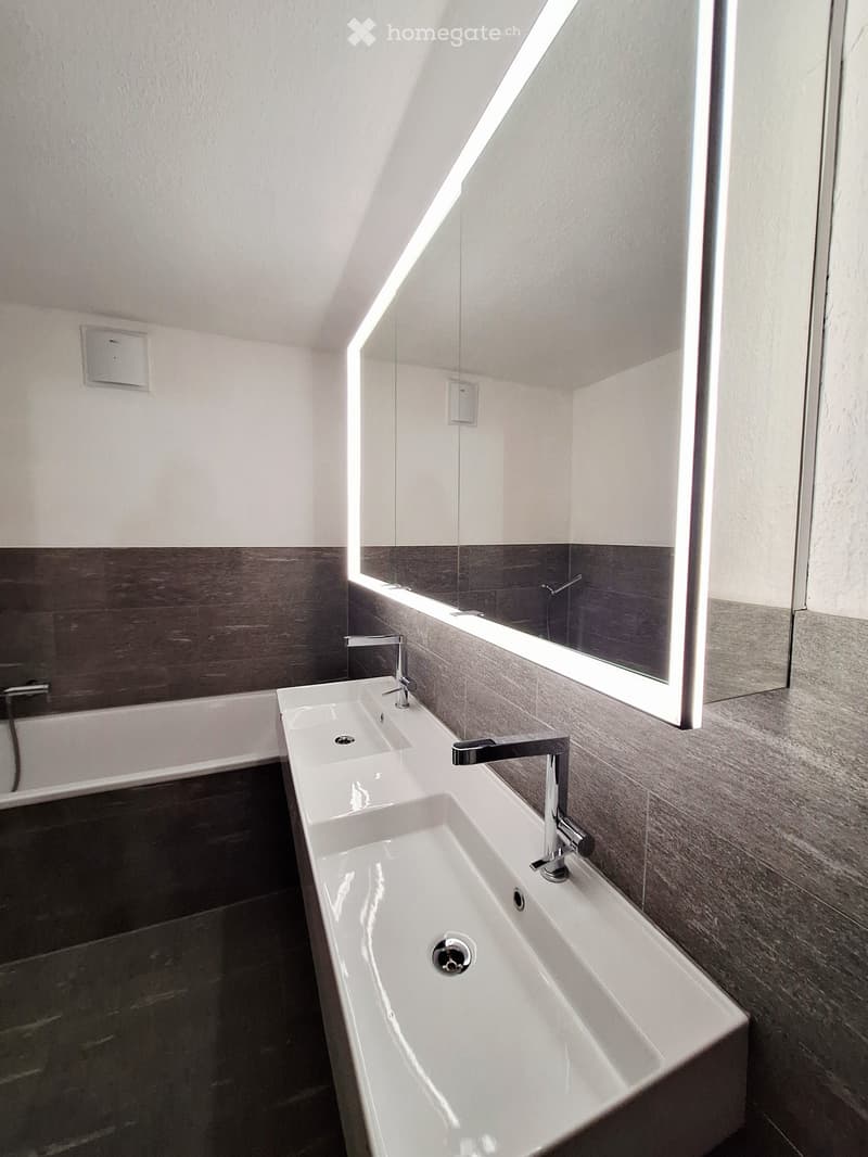 Modern bathroom with a double sink, sleek chrome faucets, a large mirror with built-in lighting, and a bathtub.