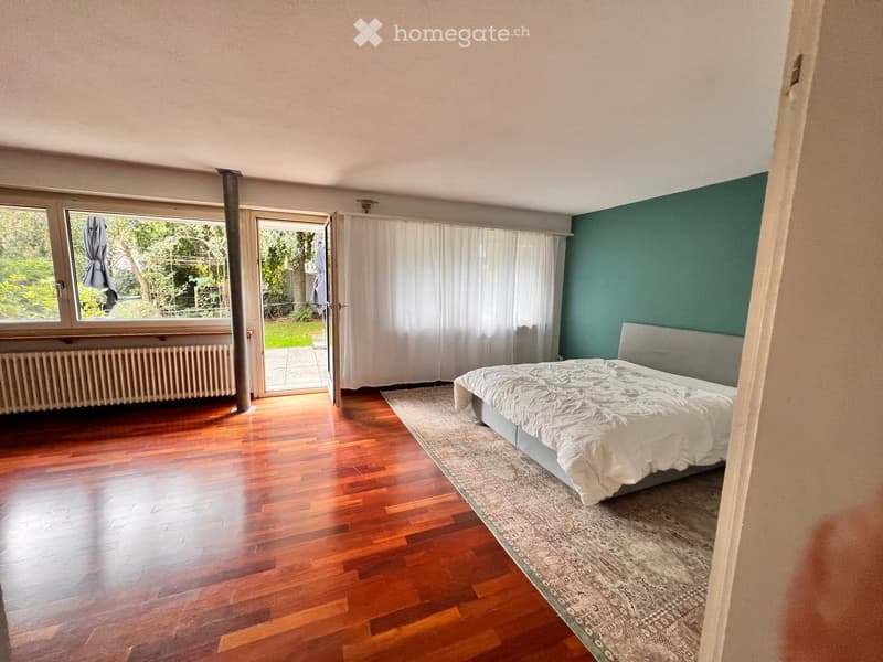 Bright bedroom with wooden flooring, a large bed with white bedding, a patterned rug, and large windows with white curtains leading to a balcony.