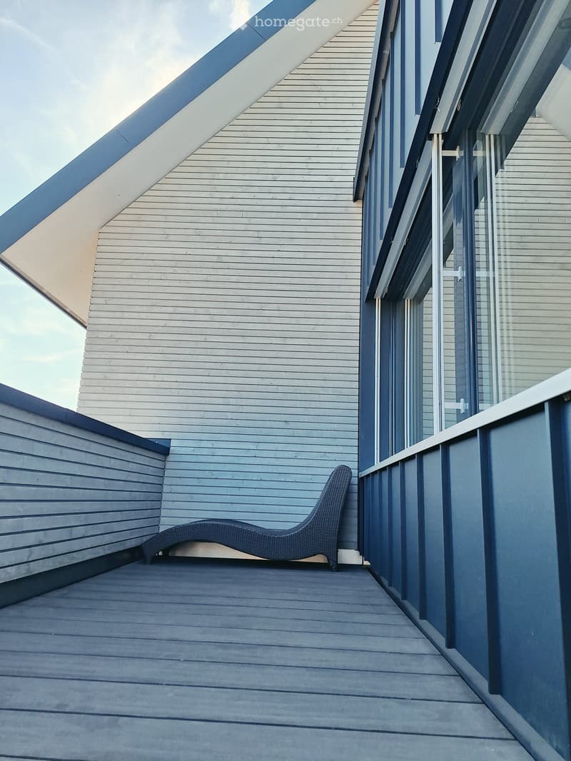 Modern balcony with wooden decking, dark blue wicker lounge chair, glass railings, and contemporary building facade.