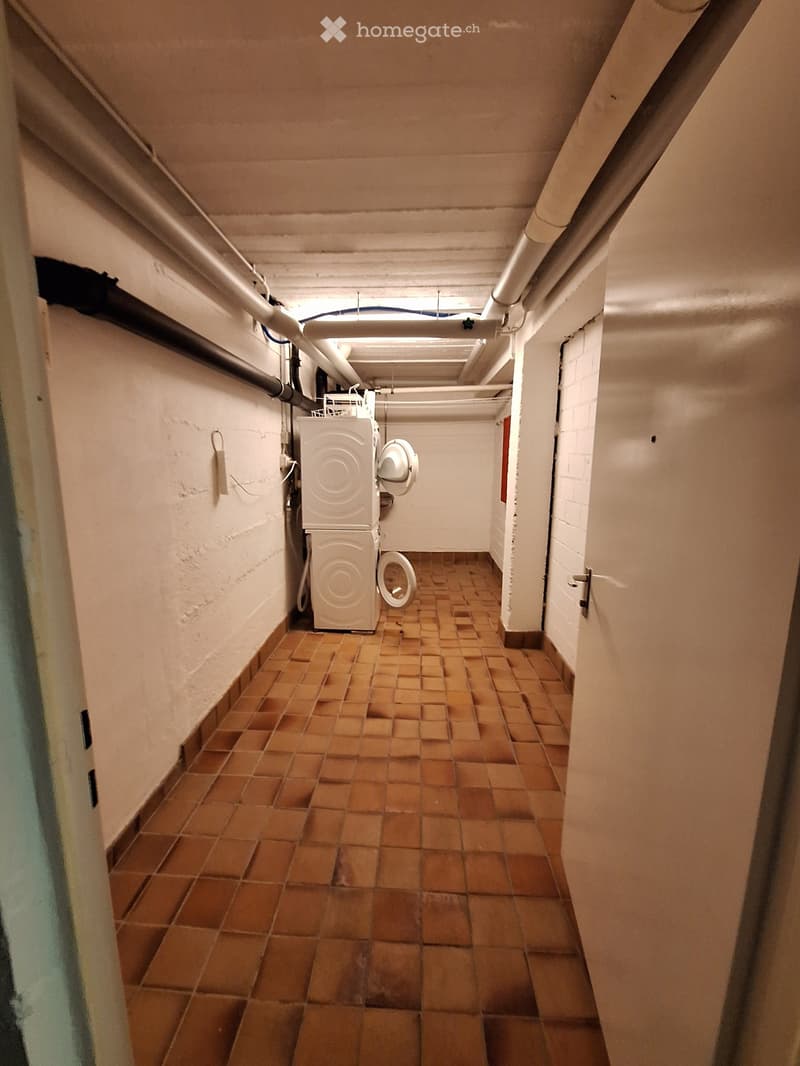 A narrow hallway with tiled flooring and white walls, featuring a washing machine and a toilet in the background.