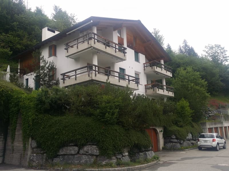A two-story house with white walls, wooden accents, multiple balconies, and a garage. Surrounded by greenery and trees.