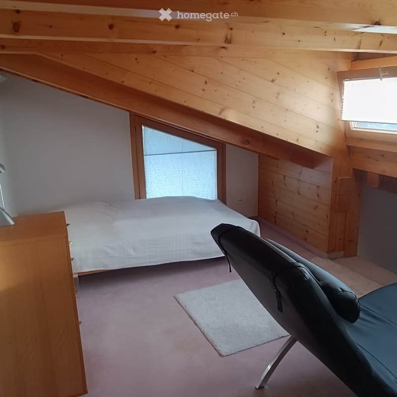 Cozy attic bedroom with wooden walls and ceiling, featuring a bed, a black leather chair, a nightstand with a lamp, and a small white rug.