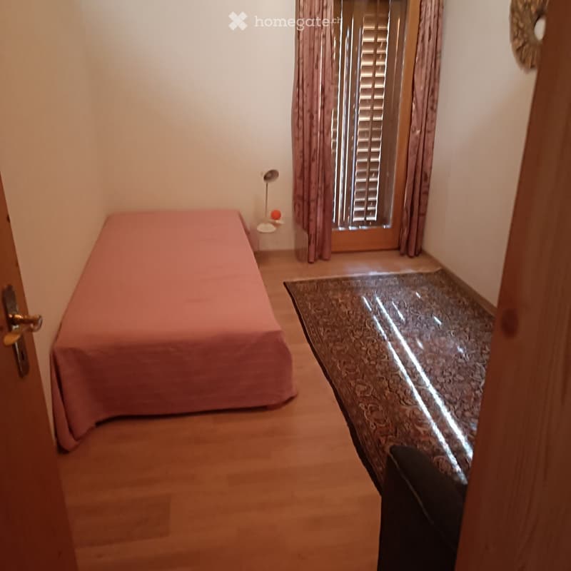 A small bedroom with a single bed, a bedside table, and a lamp near the window.