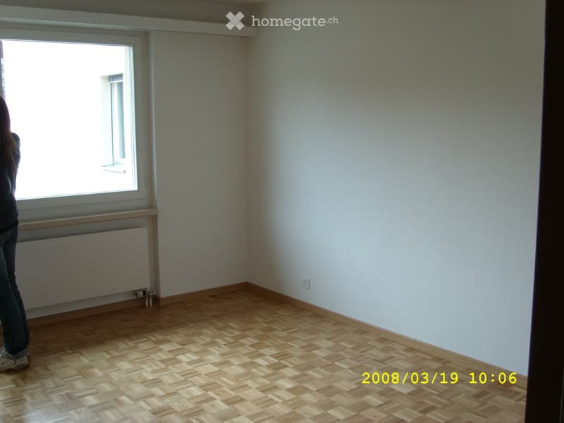 Empty room with wooden parquet flooring, white walls, and large windows allowing natural light. No furniture or furnishings are present.