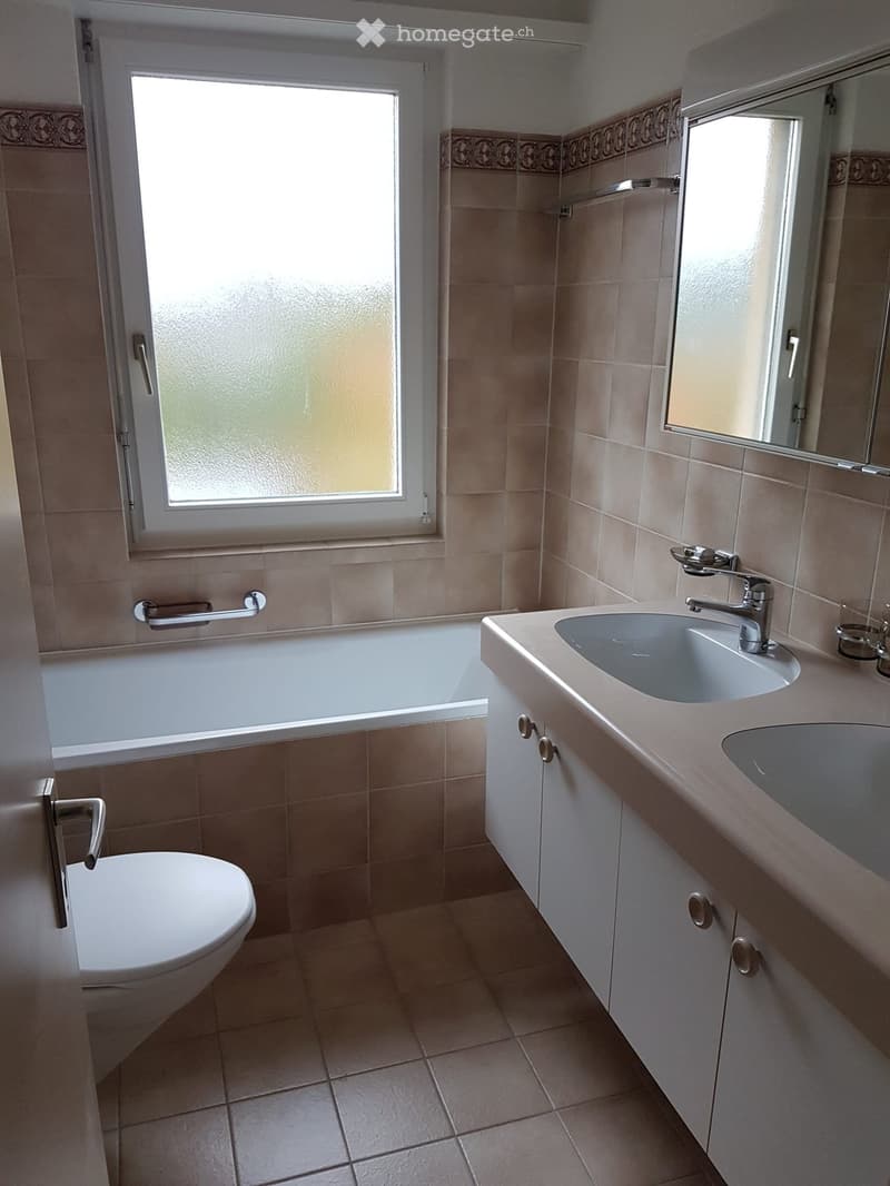 Bathroom with beige tiles, a toilet, a bathtub, a double sink with a mirror, and a window with frosted glass.